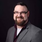 Photo of Travis Zettler, a man with dark short hair, a short beard, and glasses.