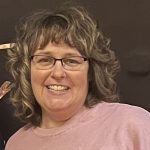 Photo of Teresa Stacey, a woman with curly dirty blonde hair and bangs, she also has glasses.
